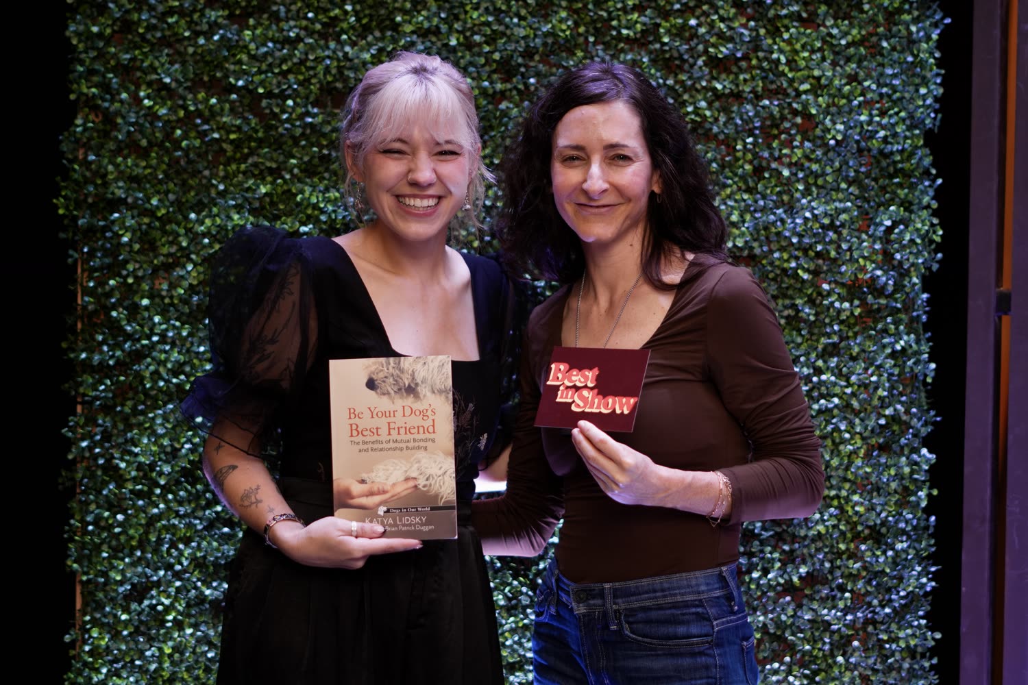 Mimi and a guest smiling in front of the green wall, holding the book Be Your Dog's Best Friend and a Best in Show card